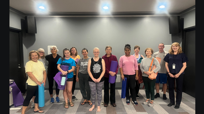 A group of people of various ages stand in a room at the Milton Public Library, holding yoga mats and posing for a group photo before or after a Free Yoga class this Summer.