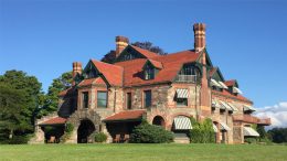 A large stone and brick mansion with multiple chimneys, red roofs, striped awnings, and arched entryways sits on a grassy lawn under a clear blue sky—home to the Milton Historical Society’s annual holiday open house.
