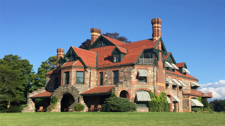 A large stone and brick mansion with multiple chimneys, red roofs, striped awnings, and arched entryways sits on a grassy lawn under a clear blue sky—home to the Milton Historical Society’s annual holiday open house.
