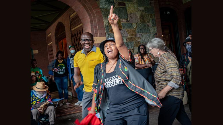 A group gathers outside a building at the Eustis Estate Summer concert; a woman in front raises two fingers while others stand and sit nearby, some smiling and wearing colorful clothing, enjoying the global rhythms of Albino Mbie.