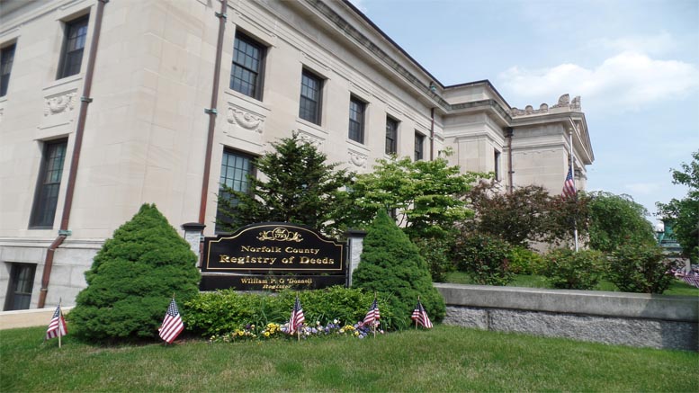 Exterior view of the Norfolk County Registry of Deeds building, with a sign and small American flags in front, surrounded by bushes and flowers—reflecting Norfolk County Real Estate activity as of April 2025.
