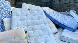 A pile of used mattresses stacked against a stone wall, showing various patterns and colors—perfect for illustrating the need for mattress disposal Milton services.