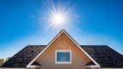 The sun shines brightly in a clear blue sky above the peaked roof of a house with a single window centered beneath the gable—summer is coming!