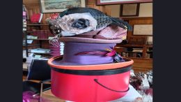A stack of vintage fashion headware in varying colors and styles is displayed on a round hat box inside a room with wooden walls and shelves of books, evoking the charming atmosphere of the Milton library.
