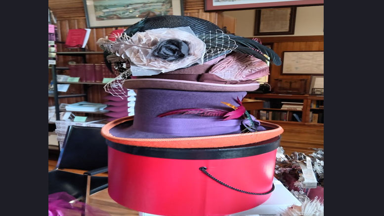 A stack of vintage fashion headware in varying colors and styles is displayed on a round hat box inside a room with wooden walls and shelves of books, evoking the charming atmosphere of the Milton library.