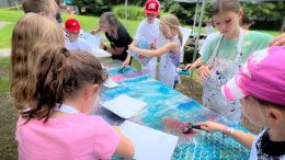 A group of children wearing aprons work on an outdoor art project, painting and pressing paper onto a bubble wrap-covered table—a scene where Summer Art Camp gives children confidence in their ability to create new worlds.
