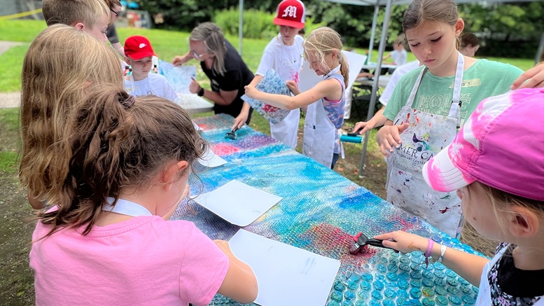 A group of children wearing aprons work on an outdoor art project, painting and pressing paper onto a bubble wrap-covered table—a scene where Summer Art Camp gives children confidence in their ability to create new worlds.