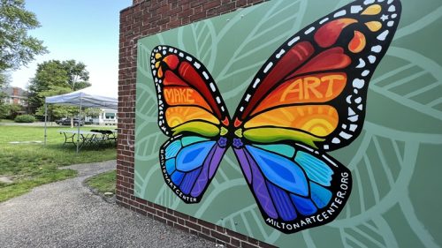 A colorful butterfly mural with the words "Make Art" on its wings brightens a brick wall near grassy tents and tables, inspiring creativity like Milton Art Center Summer Art Camp, which gives children confidence in their ability to create new worlds.