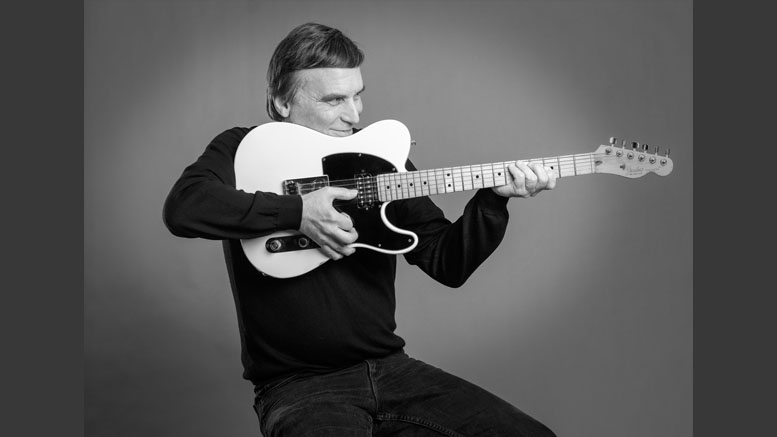 A man in a dark shirt sits holding an electric guitar horizontally, aiming it like a rifle against a plain background—an intriguing image that could headline a Wotiz Gallery photography exhibit.