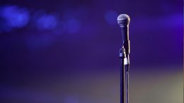 A microphone on a stand is positioned center stage against a blurred purple background, ready for the Milton Summer Concert Series featuring the Canton American Legion Band on June 25.