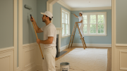 Two painters in white uniforms from Costa Painting Services add a fresh coat of paint to the light blue living room; one uses a roller, while the other works on trim from a ladder. Drop cloths cover the floor for this summer painting project.