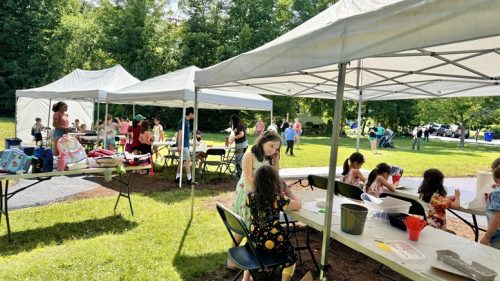 Children and adults build children confidence as they participate in arts and crafts under outdoor tents at Summer Art Camp on a sunny, grassy park day.