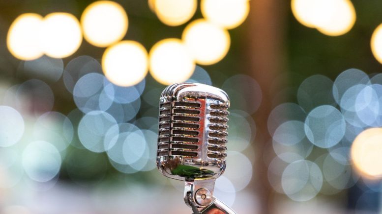 A vintage silver microphone is positioned in the foreground with blurred circular lights in the background, capturing the energy of the Milton Summer Concert Series.