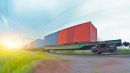 A freight train carrying shipping containers moves at speed along railroad tracks through a grassy landscape under a bright sky, highlighting the importance of avoiding common shipping container mistakes during transit.