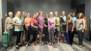 A group of women stand indoors at Milton Library Adult Yoga with Sheila Armstrong – July 22, holding yoga mats and props, smiling for a group photo on a striped floor.