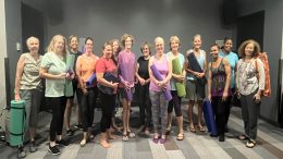 A group of women stand indoors at Milton Library Adult Yoga with Sheila Armstrong – July 22, holding yoga mats and props, smiling for a group photo on a striped floor.