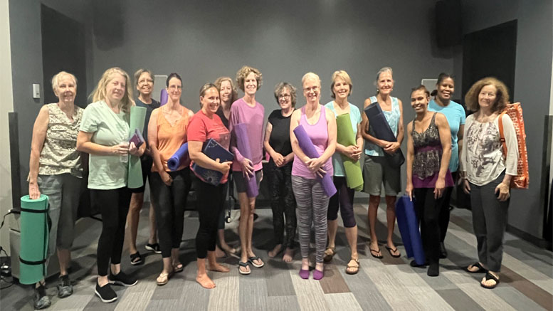 A group of women stand indoors at Milton Library Adult Yoga with Sheila Armstrong – July 22, holding yoga mats and props, smiling for a group photo on a striped floor.