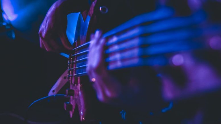 Close-up of a person playing an electric bass guitar, hands on the strings, illuminated by blue and purple stage lighting at Riverside Theatre Works starring Chuck Palmer, set to perform at Milton Summer Concert Series on July 9.