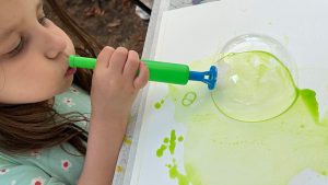 A young child, part of a junior artists art program, blows a large green bubble onto white paper with a bubble wand, creating unique green liquid stains.