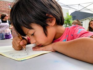 A young junior artist in a pink shirt draws intently with a pencil in a notebook while sitting at a table outdoors, perfect for any art program that encourages children to draw and paint.