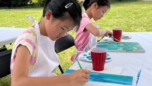 Two junior artists sit at a table outdoors, each focused on painting with brushes on canvases. They wear aprons and have red cups and paint supplies on the table.