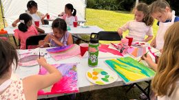 A group of junior artists sit at a table outdoors, painting colorful pictures on canvases with brushes and acrylic paints.