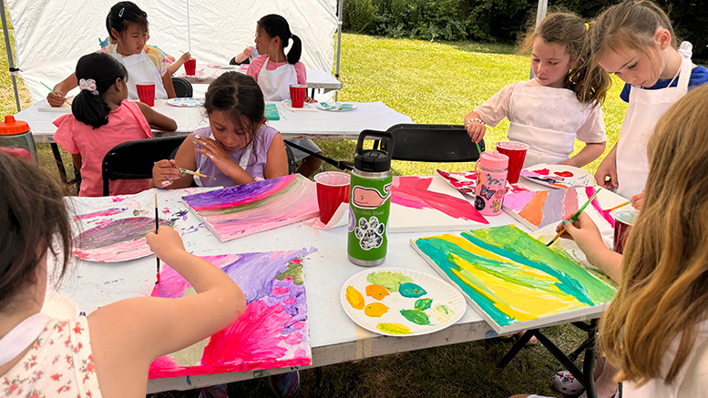 A group of junior artists sit at a table outdoors, painting colorful pictures on canvases with brushes and acrylic paints.