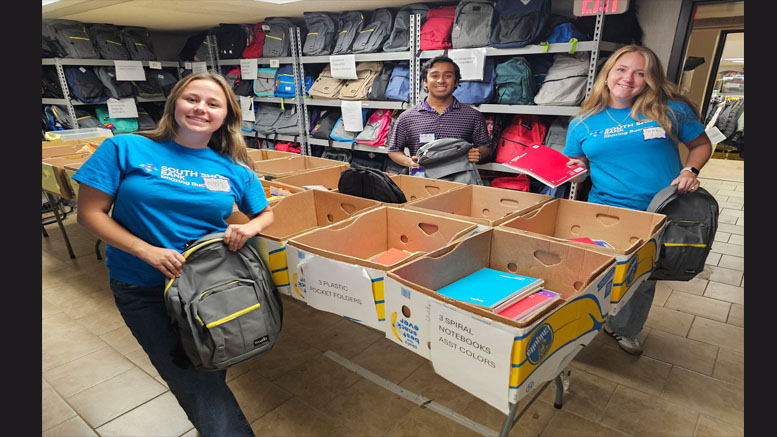 Three Interfaith volunteers stand by boxes filled with backpacks and school supplies in a storage room, preparing items for distribution to local students.