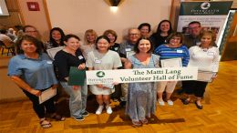 A group of people pose indoors holding a banner reading "The Matti Lang Volunteer Hall of Fame" and displaying certificates, with an Interfaith Social Services sign in the background highlighting their Halloween drive.